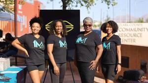 Group of Black women at a community event, dressed in black shirts featuring the blue '1M4' logo, with a red brick building and a black sign displaying a yellow logo in the background. Individuals are positioned behind a table with brochures.