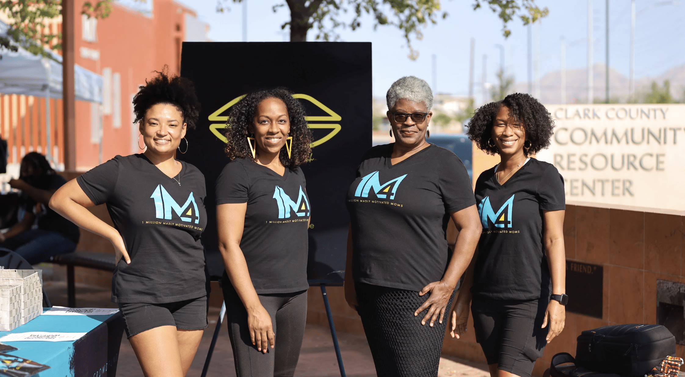 Group of Black women at a community event, dressed in black shirts featuring the blue '1M4' logo, with a red brick building and a black sign displaying a yellow logo in the background. Individuals are positioned behind a table with brochures.
