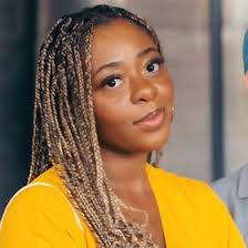Photograph of a Black woman in front of a wall with two framed pictures, wearing a blue blouse and black pants, with her right hand clenched in a fist. The framed pictures on the wall appear to be of a landscape. The background is a white wall with wooden flooring.
