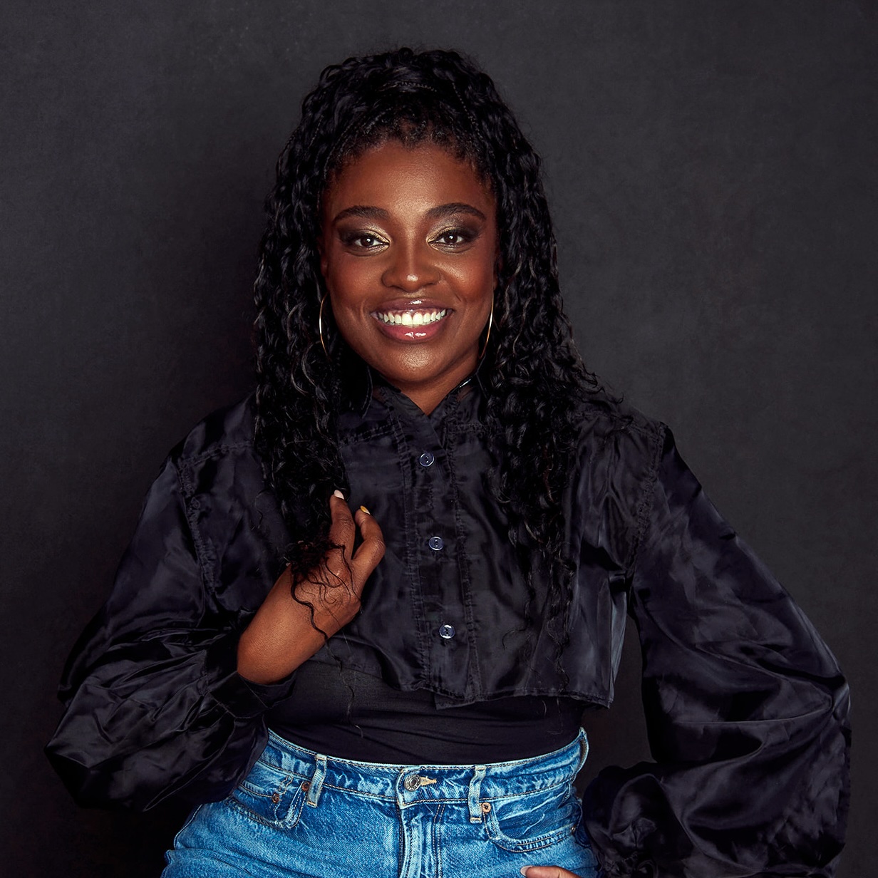 Photograph of a Black woman in front of a wall with two framed pictures, wearing a blue blouse and black pants, with her right hand clenched in a fist. The framed pictures on the wall appear to be of a landscape. The background is a white wall with wooden flooring.