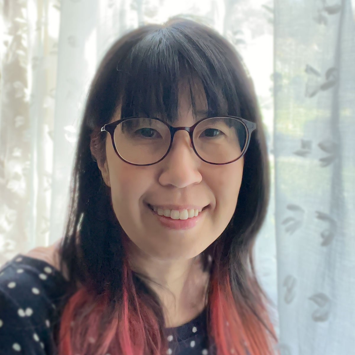 Photograph of a Black woman in front of a wall with two framed pictures, wearing a blue blouse and black pants, with her right hand clenched in a fist. The framed pictures on the wall appear to be of a landscape. The background is a white wall with wooden flooring.